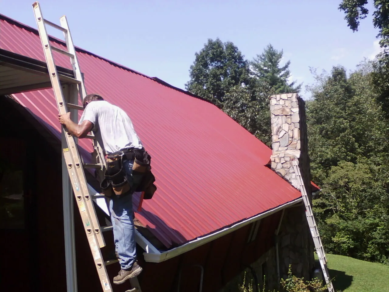 Roofer on a ladder beside a red metal roof and stone chimney — Soffit & Fascia Repair by Roofing Brothers in Steubenville